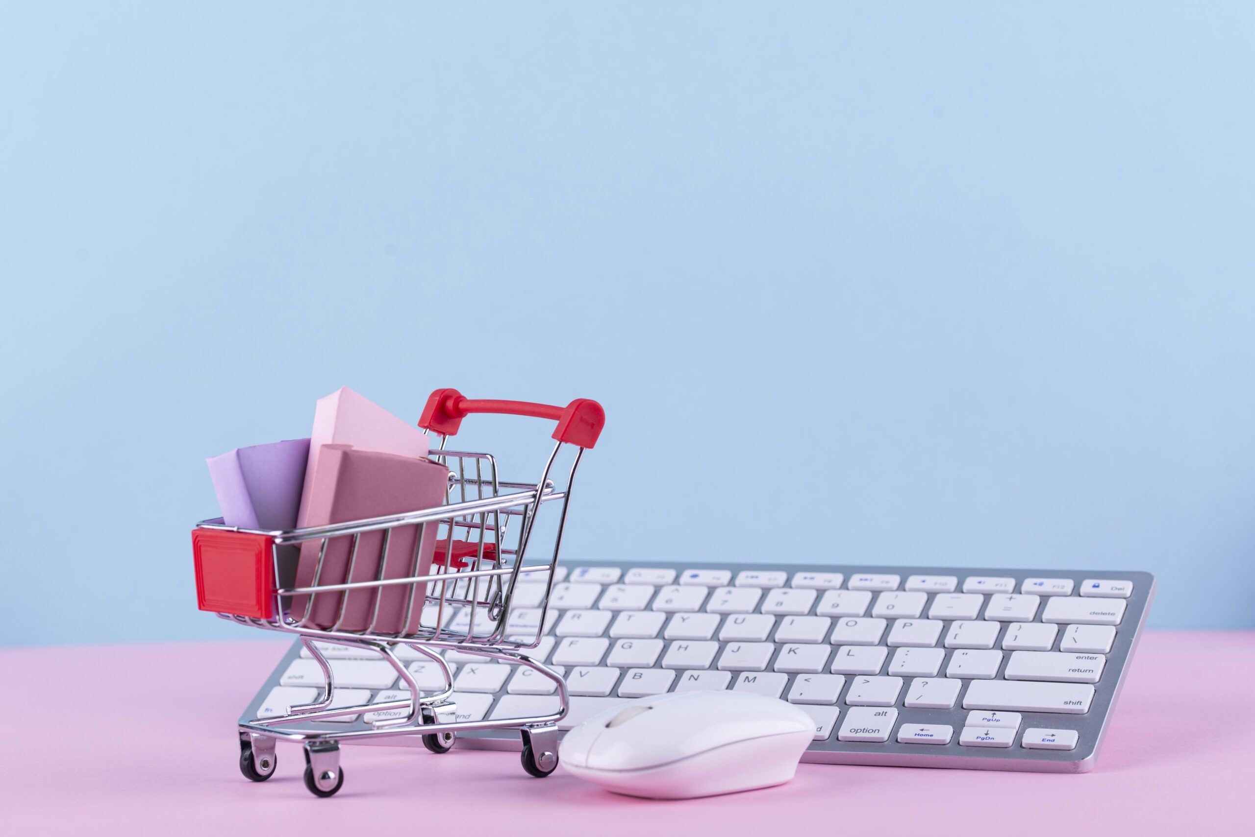 A small shopping cart with pastel boxes inside is placed next to a wireless computer mouse and keyboard on a pink and blue background.