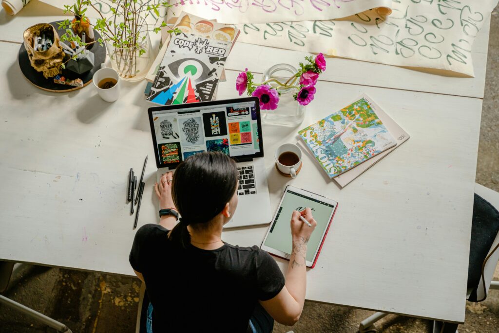 Person working at a white table with a laptop, drawing tablet, magazines, flowers in a vase, markers, and cups of coffee.