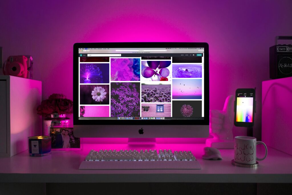 A desktop computer displays purple-themed images on the screen, surrounded by a keyboard, mouse, candle, mug, phone, and desk decor, all illuminated by pink-purple lighting.