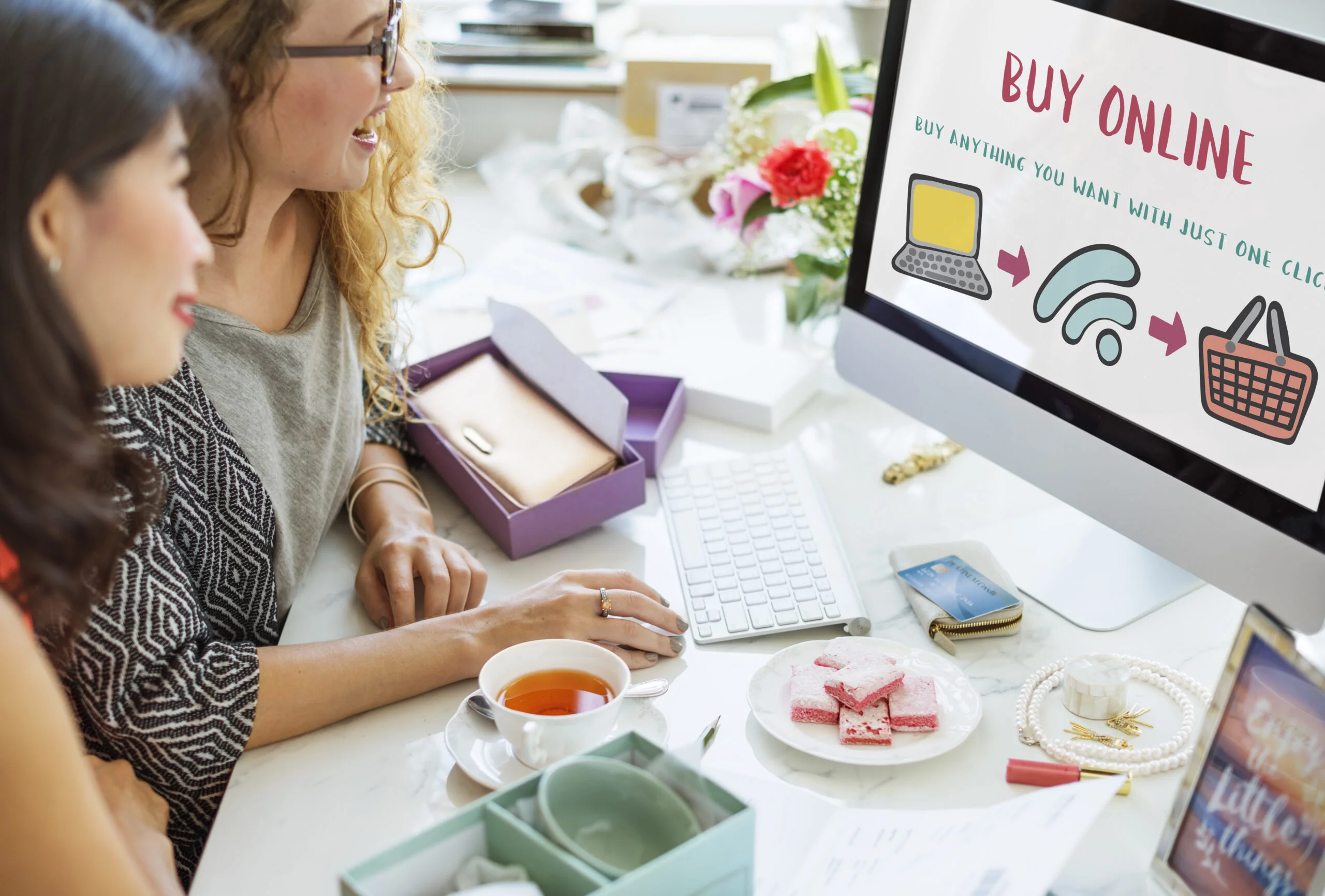 Two women smiling while shopping online, viewing a "Buy Online" screen with icons of a laptop, Wi-Fi, and a shopping basket, surrounded by tea, snacks, and packaging, representing e-commerce website content writing.
